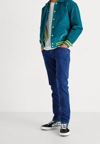 Teal jacka med gula knappar och randiga ärmar, vit skjorta, blå jeans och svarta rutiga sneakers. Avslappnad outfit med lagrade texturer.