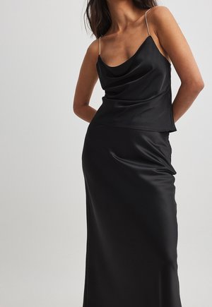 Kvinde iført en sort satin natkjole med rhinsten tynde stropper, poserende med hænderne bag ryggen mod en ensartet lys baggrund.