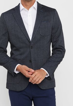 Mand iført en mørkegrå fiskegratsmønster blazer med en hvid skjorte og marineblå bukser, hænderne foldet foran.