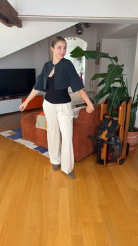 Jeune femme debout dans un salon avec un sol en bois, portant un haut noir et un pantalon blanc, près de plantes et d'un appareil d'exercice plié.