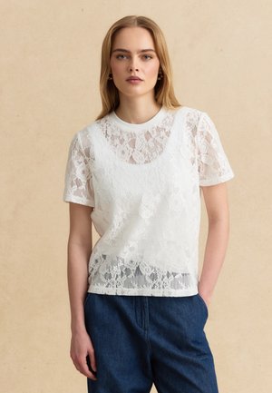 Jonge vrouw draagt een wit kort mouwtopje van bloemrijke kant over een wit ondershirt, gecombineerd met loszittende donkerblauwe broek.