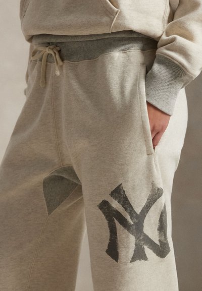 Grå joggebukser i en lys beige tone, med en fremtredende logo i svart på venstre ben og et strukturert belte med snøring.