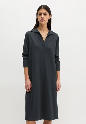 Femme portant une robe gris foncé à manches longues, longueur genoux, avec un col et un décolleté en V, debout devant un fond blanc uni.