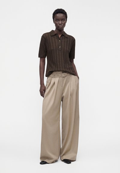 Femme portant un polo à manches courtes brun foncé et un pantalon beige taille haute à jambes larges avec ceinture, debout avec une main dans la poche.