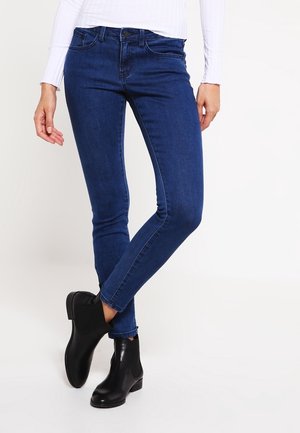 Person, die dunkelblaue Skinny-Jeans, schwarze Knöchelstiefel und ein weißes langärmliges Oberteil trägt, steht mit überkreuzten Beinen.