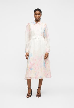 Kvinde iført en hvid midi-kjole med lange ærmer med pastelfarvede blomster, bælte i taljen og sorte sandaler med høje hæle og stropper.