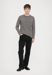 Grå langermet skjorte med "A|X ARMANI EXCHANGE" logo, kombinert med svarte rett ben jeans og svarte sko. Glatt stoff, avslappet passform.