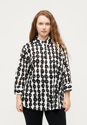 Femme aux longs cheveux roux portant un pantalon noir et une chemise boutonnée à motifs noir et blanc, debout devant un arrière-plan clair uni.