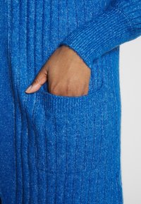 Cardigan tricoté bleu avec des côtes verticales, des manches longues et une poche avant. La texture semble douce et chaude.