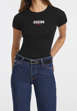 Camiseta negra de rayas con mangas cortas, con el logo "GUESS JEANS" en blanco y rojo. Combinada con jeans de mezclilla azul oscuro de tiro alto y un cinturón negro.