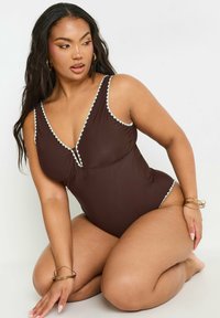Monokini marrón con escote en V profundo, tejido suave, ribete azul claro en contraste y cobertura moderada. La modelo posa de rodillas.