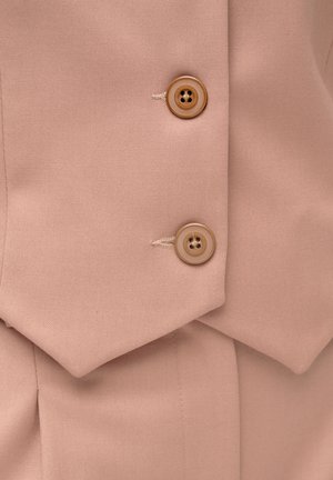 Zbliżenie na beżową tkaninę z dwoma jasno-brązowymi guzikami przyszytymi na nakładających się klapach dopasowanego ubrania.