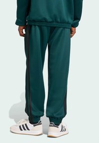 Pantalon de survêtement teal avec des rayures latérales noires, des poignets élastiques et une taille avec cordon de serrage, associé à des baskets blanches et noires.