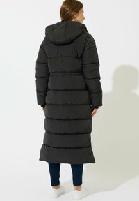 Kvinde iført en lang sort hættetrøje, blå jeans og hvide sneakers, stående med ryggen til på en hvid baggrund.