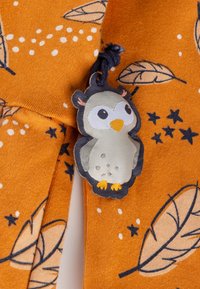 Oranger Stoff mit einem Blatt- und Sternmuster, an dem ein graues und gelbes Eulenschild mit einer dunkelblauen Schnur befestigt ist.