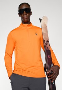 Oranges Langarm-Pullover mit einem Viertelreißverschluss und einem Spinnenlogo. Das Model hält Skier und trägt schwarze Skibrille. Graue Hose rundet das Outfit ab.
