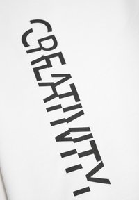 Witte t-shirt met het woord "CREATIV4TY" in vetgedrukte, zwarte sans-serif typografie in een diagonale positie over de stof.