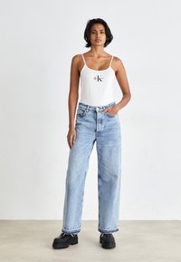 Hvit tanktop med logo, kombinert med lyseblå vide jeans med rivet nederst, og svarte sko med tykk såle. Enkelt, uformelt antrekk.
