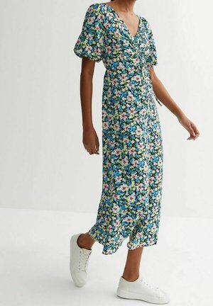 Vrouw draagt een blauwe midi-jurk met bloemenprint, pofmouwen en witte sneakers, lopend op een effen witte achtergrond.