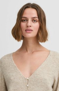 Beige neulecardigan V-pääntiellä, ribatoidu pintastruktuurilla, ja kolme helmeä edessä. Pehmeä, mukava materiaali herkällä yksityiskohdalla.