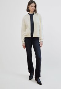 Chaqueta bomber color crema con textura moteada, puños y cuello de canalé; combinada con vaqueros negros acampanados y botines negros.