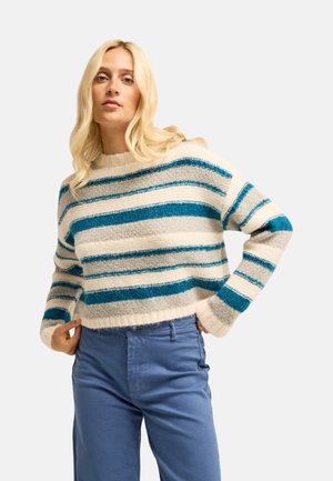 Donna con lunghi capelli biondi che indossa un maglione a righe bianche, grigie e blu e pantaloni blu su uno sfondo chiaro uniforme.