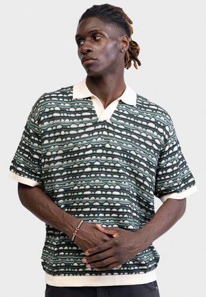 Άνδρας με ντραντλόκς που φοράει κοντομάνικο πλεκτό πουκάμισο με σχέδια, λευκό γιακά και μανσέτες, τα χέρια του είναι σταυρωμένα μπροστά, κοιτάζει προς τα πλάγια.