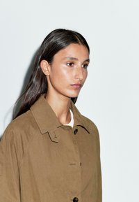 Bruine lichtgewicht jas met een kraag, knoopsluiting en een gladde textuur. Model heeft lang, donker haar en draagt een simpel wit topje eronder.