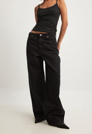 Frau trägt schwarze Jeans mit weitem Bein, schwarzes Spaghettiträger-Oberteil und schwarze spitze Schuhe, steht vor einem schlichten hellen Hintergrund.