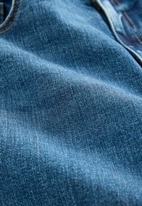Denimstof in een medium wassing blauw, met een subtiele textuur en een zichtbaar weefpatroon. Close-up toont de zachtheid en een lichte glans.