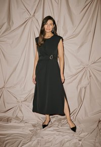 Čierne bezrukávové šaty so bočným rozparkom, s opaskom na páse s oválnou sponou. Hladká tkanina na pokrčenej, jemne ružovej pozadí.