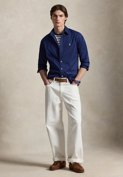 Νέος άνδρας που φοράει μπλε-σκούρο πουκάμισο πάνω από ριγέ μπλουζάκι, λευκό παντελόνι με πλεκτή ζώνη, καφέ παπούτσια τύπου μπότα και ρολόι χειρός, στέκεται με τα χέρια στις τσέπες.