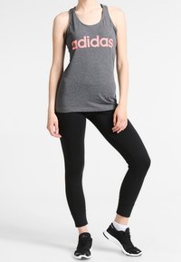 Vrouw in een grijze Adidas sporttop, zwarte leggings, witte sokken en zwarte sportschoenen, staand met één been licht gebogen.