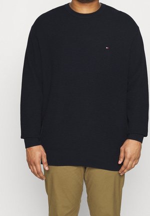 Mann trägt einen schwarzen, strukturierten Langarmpullover mit einem kleinen rot-weiß-blauen Logo auf der linken Brustseite und Khakihosen.