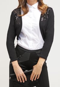 Donna che indossa una camicia bianca con bottoni e un cardigan nero cropped con dettagli in paillettes, tiene una borsa a mano nera con entrambe le mani.
