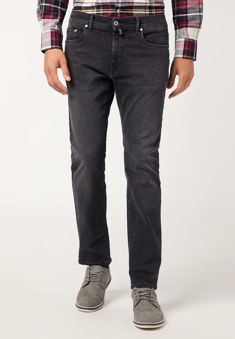 Jeans neri da uomo in denim con vestibilità slim, design a cinque tasche, chiusura con bottone e una sottile texture scolorita, abbinati a scarpe grigie.