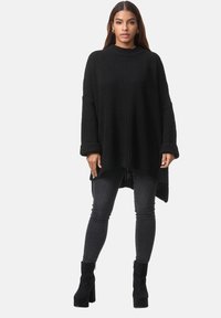 Pullover nero oversize in maglia con maniche larghe, texture a coste e orlo asimmetrico, abbinato a jeans skinny scuri e stivaletti neri con tacco.