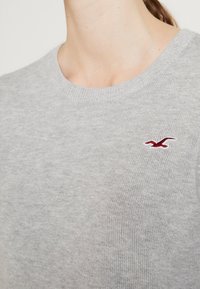 Hellgrauer Strickpullover mit Rundhalsausschnitt, der ein kleines rotes Vogel-Logo auf der linken Brust verfügt. Weiche Textur mit gerippten Kragenrändern.