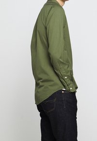 Långärmad olivgrön skjorta med krage som knäpps, av bomullstyg och med rundad fåll, bärs med mörka jeans i denim och synliga sömnadsdetaljer.