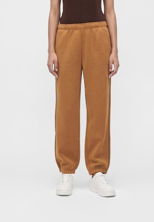 Personne portant un pantalon de jogging marron à taille élastique avec coutures latérales et des baskets blanches, debout devant un fond clair uni.