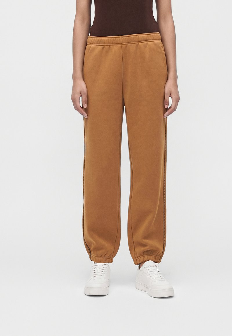 Personne portant un pantalon de jogging marron à taille élastique avec coutures latérales et des baskets blanches, debout devant un fond clair uni.