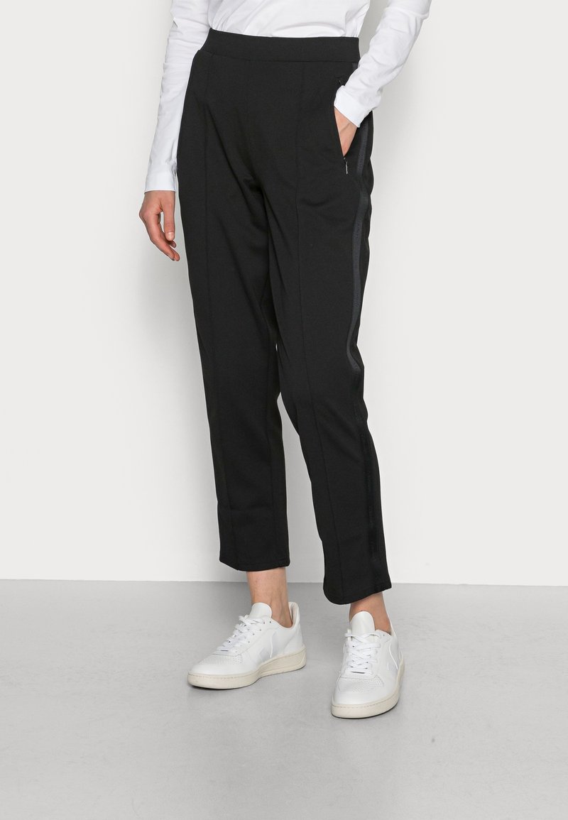 Zwarte tapered broek met een gladde textuur, elastische tailleband en zijzakken. Gecombineerd met witte sneakers voor contrast.