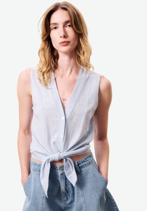 Femme aux cheveux blonds mi-longs portant un haut bleu clair sans manches boutonné et noué à la taille, avec un pantalon bleu texturé, les mains dans les poches.