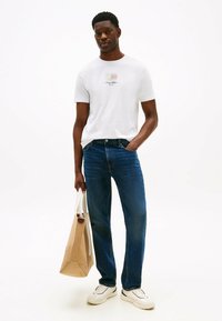 Hvid t-shirt med kort ærme og logo, parret med blå denim jeans og hvide sneakers, mens der bæres en beige tote bag. Rent, afslappet look.