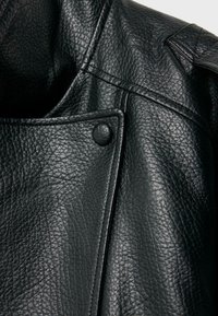 Close-up of textured black leather jacket with stitched seams, a snap button, and part of the collar visible on a white background.