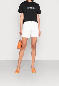 Black cropped T-shirt with white logo, paired with white shorts. Accessorized with an orange patent handbag and orange slide sandals.