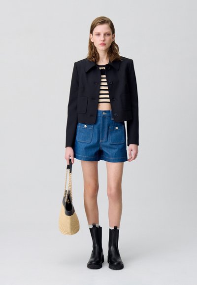 Chaqueta negra corta con botones grandes, camiseta de rayas, pantalones cortos de mezclilla, botines negros y un bolso de paja con un asa negra.