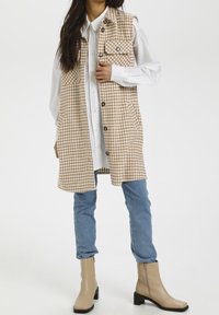 Gilet lungo a scacchi marrone e beige con tasche, indossato sopra una camicia bianca, abbinato a jeans blu e stivaletti beige.