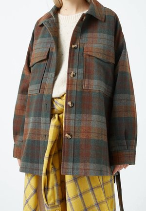 Femme portant une veste à boutons à carreaux marron, vert et gris sur un pull crème, associée à une jupe à carreaux jaune et violet nouée.