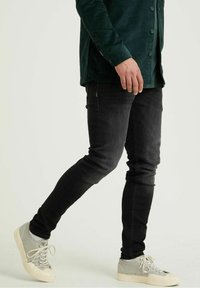 Jeans ajustados negros, combinados con zapatillas gris claro. El conjunto incluye una camisa de pana verde oscuro con detalles de botones y tejido texturizado.